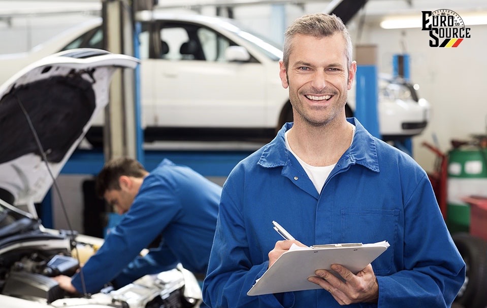 Automotive technician holding a clipboard smiling with another technician in the background working on a car.